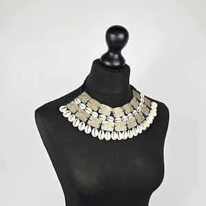 May include: A multi-layered statement necklace featuring rows of off-white cowrie shells and square, etched silver-toned metal plates. The design is arranged in a wide, collar-style formation with a black cord base, creating a textured, layered appearance.