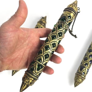 May include: A cylindrical metal case with an antique brass finish, featuring a repeating scale-like pattern with inlaid dark green glass accents. The object has pointed ends, a screw-top cap, and a small metal chain attached to the top section.