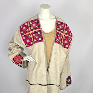 May include: A light beige open-front jacket with colourful embroidered floral patterns on the shoulders and sleeves. The jacket has a gold-coloured lining and a decorative trim along the edges. The embroidery features pink, yellow, and green accents.