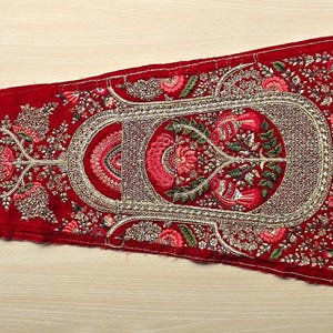Puede incluir: Un panel de tela de terciopelo rojo con bordados intrincados que presentan motivos florales y de árboles en hilo plateado y dorado. El panel tiene un borde festoneado y probablemente forma parte de una prenda tradicional india.