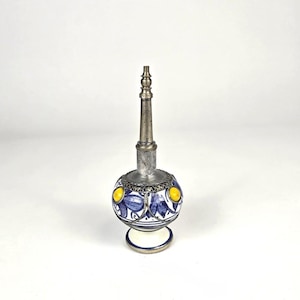 May include: A decorative perfume bottle with a silver metal top and base. The round ceramic body is white with blue floral designs and yellow accents. The bottle stands approximately 6 inches tall.