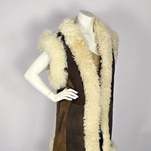 Può includere: Un lungo gilet in pelle scamosciata marrone con collo e fodera in pelliccia di pecora bianca. Il gilet ha un collo a scialle e una silhouette lunga e fluida.
