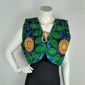 May include: A navy blue vest with green and gold embroidery. The vest features circular and floral patterns, with mirrored accents. The edges are trimmed in green, and the vest has a gold-colored tie closure.
