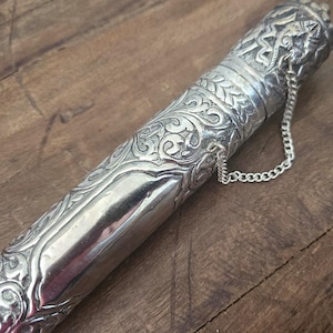May include: Close-up of a silver metal object with intricate floral and geometric patterns. The object has a cylindrical shape with a decorative cap and a chain attached. The surface reflects light, highlighting the detailed engravings. The item appears to be a decorative container or sheath.