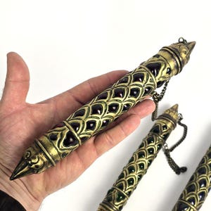 May include: A cylindrical metal case with a gold-toned finish featuring an intricate scale pattern and embedded dark red stones. The object has a pointed cap at each end and is attached to a small metal chain, showcasing a detailed relief design.