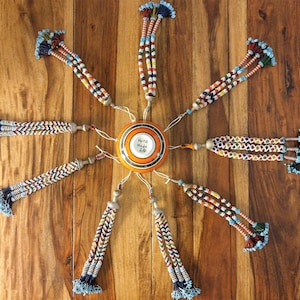 Op de afbeelding: Een bovenaanzicht van een decoratief stuk met kralenkwasten die uitstralen van een centrale oranje en witte schijf. De kwasten zijn veelkleurig met blauwe, rode, gele en witte kralen. Op de schijf staat "Hand Made 26". De achtergrond is een houten oppervlak.