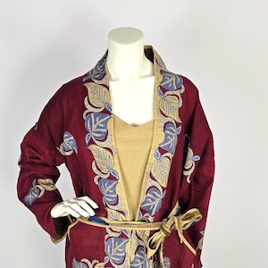 Può includere: Un accappatoio bordeaux con ricami a foglie blu e oro. L'accappatoio ha una fodera beige e una cintura abbinata. L'accappatoio è esposto su un manichino. È un capo d'abbigliamento unico.