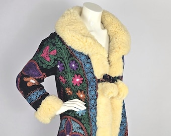 May include: A black coat with colorful floral embroidery and a cream-colored shearling collar and cuffs. The coat features a decorative belt with pom-poms and is displayed on a mannequin. The embroidery includes pink, purple, green, and orange flowers.