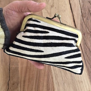 May include: A small, black and white zebra-striped coin purse with a brass clasp. The purse is held open, revealing the interior. The background is a wooden surface.