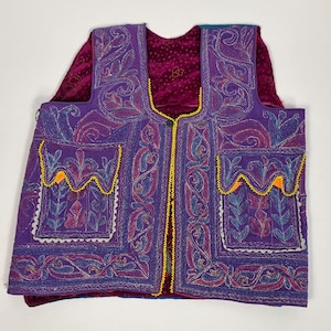 May include: A purple embroidered vest with intricate floral and paisley patterns. The vest features two front pockets with yellow and orange trim, and a yellow beaded trim along the edges. The vest is layered over a burgundy velvet top.