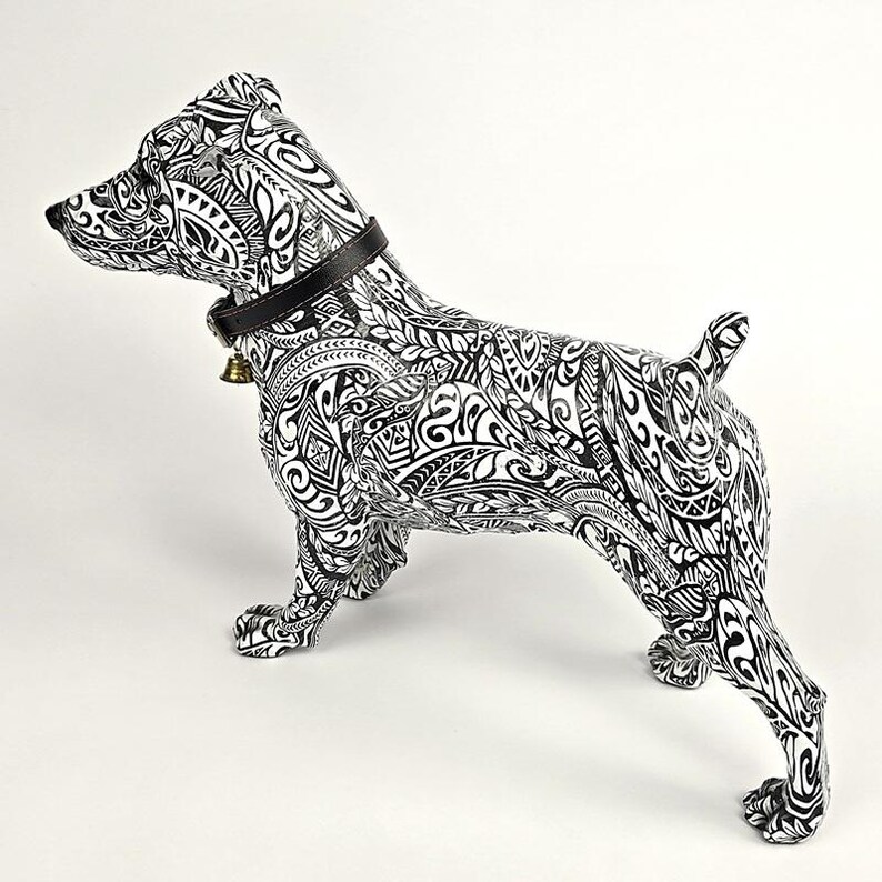 May include: A decorative dog figurine in a standing pose, featuring a black and white tribal pattern. The dog wears a black collar with a small gold bell. The figurine is set against a white background.
