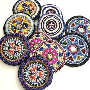 Könnte beinhalten: Eine Sammlung farbenfroher, bestickter Untersetzer. Die Untersetzer weisen komplizierte Muster in verschiedenen Farben auf, darunter Blau, Rot, Gelb und Weiß. Einige Untersetzer haben verspiegelte Zentren, andere einfarbige Designs. Die Untersetzer sind rund und haben einen schwarzen Rand. Maße in Zentimetern.
