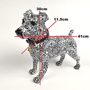 May include: A black and white decorative dog statue with intricate patterns. The statue measures 16 inches long, 12 inches high, and 4.5 inches wide. It features a black collar with a gold bell.