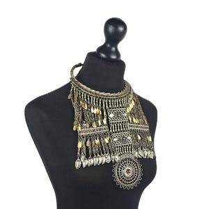 Puede incluir: Un collar tipo pechera de varias capas en acabado de latón antiguo con un collar trenzado, dijes colgantes en forma de hoja, paneles rectangulares con pequeñas piedras incrustadas de colores y un gran colgante circular de filigrana.