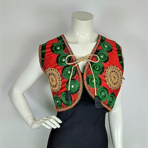 May include: A sleeveless vest with a vibrant red background, adorned with green circular patterns and gold embroidered details. The vest features a tie closure and black tassels. The garment is displayed on a mannequin.