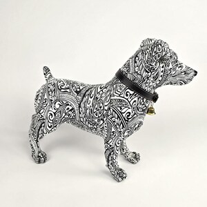 May include: A decorative dog figurine with a black and white tribal pattern. The dog has a black collar with a small gold bell. The figurine is standing on all fours, with its tail up and head turned to the right.
