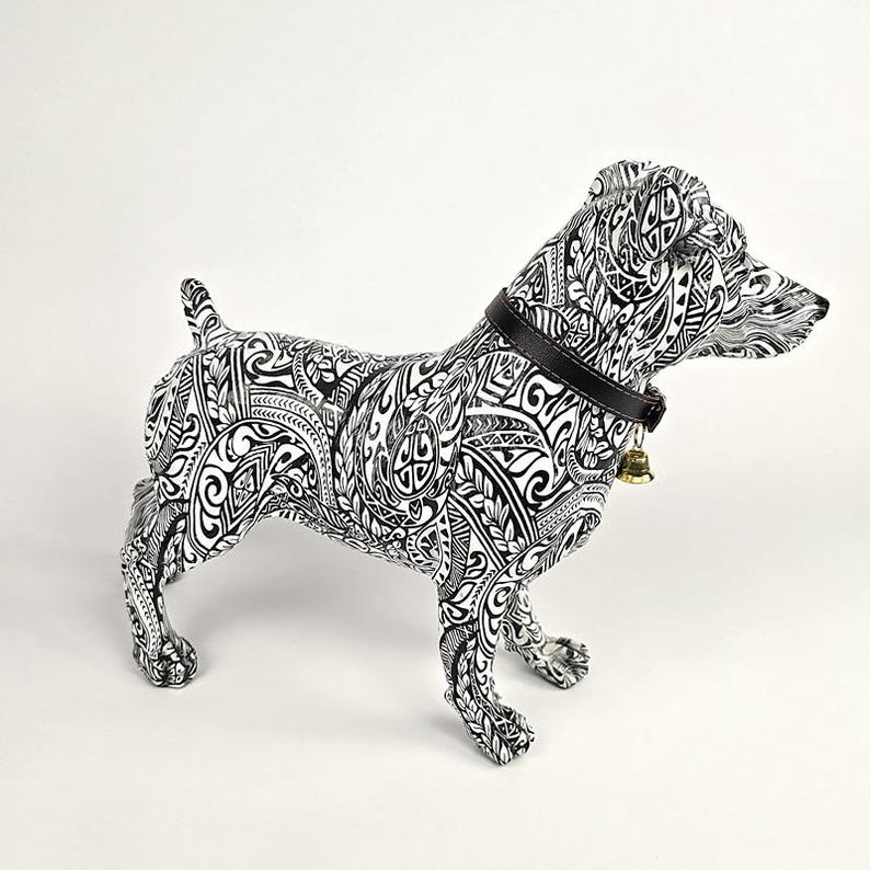 May include: A decorative dog figurine with a black and white tribal pattern. The dog has a black collar with a small gold bell. The figurine is standing on all fours, with its tail up and head turned to the right.