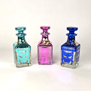 May include: Three decorative glass bottles in teal, pink, and blue. Each bottle has a square shape with a matching stopper. The bottles feature ornate silver designs and floral accents, creating a vintage aesthetic.