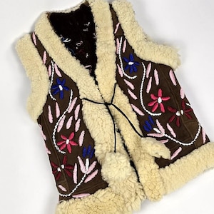 Könnte beinhalten: Eine braune Wildlederweste mit einem weißen Lammfellfutter. Die Weste ist mit rosa, roten und blauen Blumen bestickt. Die Weste hat einen Bindeverschluss am Hals.