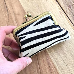 Peut inclure: Un petit porte-monnaie zébré avec un fermoir en laiton. Le sac à main présente un motif noir et blanc et un cadre en laiton. Le fermoir est de forme sphérique. Le sac est tenu dans la main d'une personne.