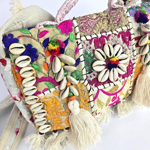 Op de afbeelding: Een kleurrijke patchwork handtas met gedetailleerde borduursels, schelpen en kwastjes. De tas heeft levendige bloempatronen in roze, blauw en geel, met een gevlochten handvat. Het ontwerp bevat verschillende texturen en versieringen voor een bohemian uitstraling.