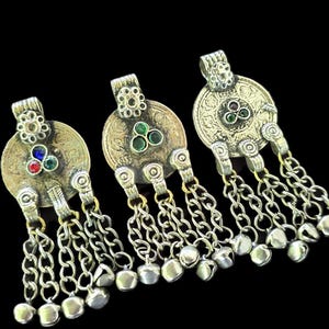 May include: Three silver-toned metal pendants featuring circular discs with embossed patterns, a floral-shaped top bail, and a central cluster of three small round stones. Each pendant has five dangling silver chains ending in small metal bells.
