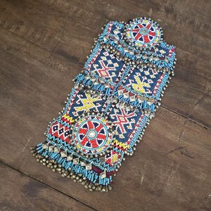Puede incluir: Textil adornado con cuentas en tonos azules, rojos, amarillos y plateados. El intrincado diseño presenta patrones geométricos y motivos circulares, con una franja de pequeñas campanas plateadas. La pieza se exhibe sobre una superficie de madera.