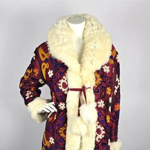 Puede incluir: Una larga chaqueta de color burdeos con cuello y puños de piel blanca. La chaqueta está bordada con coloridos patrones florales y geométricos.