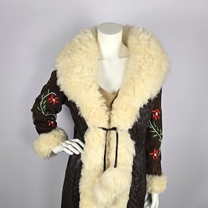 Può includere: Un lungo cappotto in pelle scamosciata marrone con colletto e fodera in montone bianco. Il cappotto ha fiori ricamati sulle maniche e una cintura in vita.