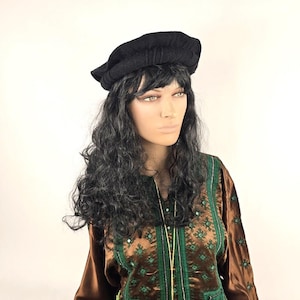 Può includere: Un cappello nero di feltro con un bordo ripiegato, indossato da una persona con lunghi capelli neri ricci. La persona indossa una camicia con un motivo marrone e verde.