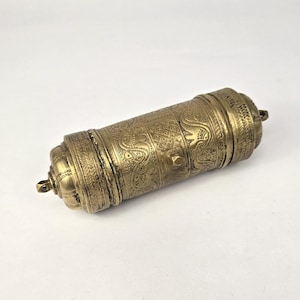 Puede incluir: Un recipiente cilíndrico de latón antiguo con intrincados patrones en relieve. El recipiente presenta diseños detallados, incluidos motivos de elefantes, y está rematado en ambos extremos con pináculos decorativos. El color general es un latón cálido y envejecido.