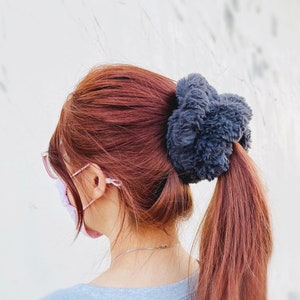 May include: A woman with long, auburn hair tied back with a fluffy, gray scrunchie. She is wearing a light gray sweater and a pink face mask.