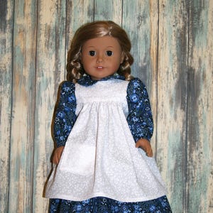 May include: A doll with light brown hair styled in two braids, wearing a long-sleeved dress featuring a dark blue floral pattern. The dress includes a white pinafore overlay with a small pocket on the front, a rounded collar, and dark brown shoes.