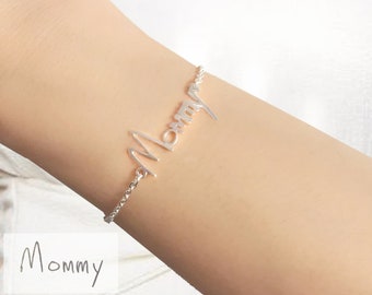 May include: Silver bracelet featuring the word "Mommy" in cursive script. The bracelet is worn on a person's wrist.