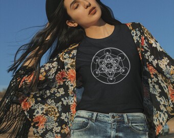 May include: A woman wearing a black t-shirt with a white geometric design, a floral kimono, and denim shorts. She is wearing a black hat and has her hair pulled back.