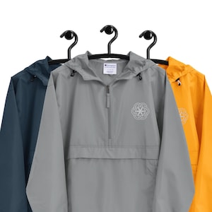 Puede incluir: Tres cortavientos colgados en perchas. El cortavientos del centro es gris con un logotipo blanco en el pecho izquierdo. El cortavientos de la izquierda es azul marino y el cortavientos de la derecha es amarillo.