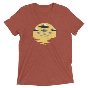 Puede incluir: Una camiseta roja con un gr&aacute;fico de un sol amarillo y m&uacute;ltiples platillos voladores en formaci&oacute;n.