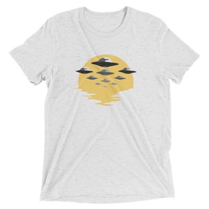 Puede incluir: Una camiseta blanca con un gr&aacute;fico de un sol amarillo con un reflejo en el agua y varios OVNIs grises volando sobre &eacute;l.