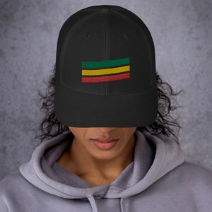 Puede incluir: Una gorra de camionero de malla negra con un dise&ntilde;o de rayas horizontales verdes, amarillas y rojas. La gorra tiene una visera curvada y una parte trasera de malla.