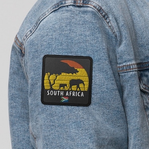 May include: A light blue denim jacket with a sherpa collar and a patch on the left sleeve. The patch features a sunset scene with elephants and the text "SOUTH AFRICA" with the South African flag.