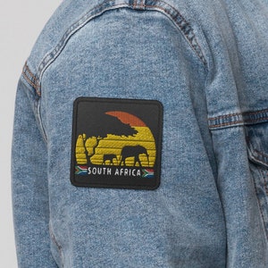 Può includere: Una giacca di jeans blu chiaro con colletto in sherpa e una toppa sulla manica sinistra. La toppa raffigura un tramonto con un albero, animali e il testo "Sudafrica".