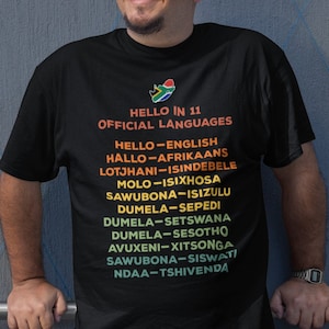 May include: Black t-shirt with the text "HELLO IN 11 OFFICIAL LANGUAGES" and a South African flag graphic. Below the text are the words "HELLO-ENGLISH" and other languages. The person is wearing blue jeans.