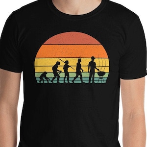 May include: A black t-shirt with a vintage-style graphic of the evolution of man, culminating in a person grilling. The graphic features a sunset with orange, yellow, and blue stripes.
