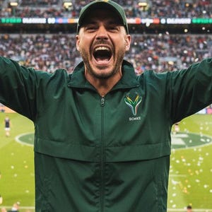Puede incluir: Un hombre con chaqueta y gorra verdes celebra con los brazos en alto en un estadio. La chaqueta tiene un logo y la palabra "BOKKE". El fondo muestra una multitud y un campo deportivo.