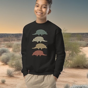 Op de afbeelding: Een jong persoon draagt een zwart sweatshirt met een retro-ontwerp van vier gordeldieren in verschillende kleuren. Het sweatshirt heeft een ronde hals en lange mouwen. De persoon staat in een woestijnsetting met een echt gordeldier op de voorgrond.