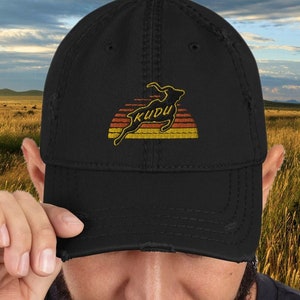 May include: A black distressed baseball cap with a vintage-style embroidered design of a kudu antelope jumping over a sunset. The text "KUDU" is embroidered in yellow on the cap.