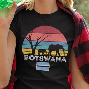 May include: A woman wearing a black t-shirt with a retro sunset graphic and the text "BOTSWANA" sits on a red and black plaid blanket. The graphic features two elephants and a tree silhouette. She is holding a green water bottle and an orange.