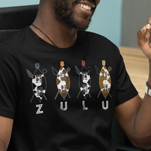 May include: A black t-shirt featuring a graphic of four shields and spears, with the word "ZULU" printed below. The shields are decorated with a black and white pattern and have colourful accents.