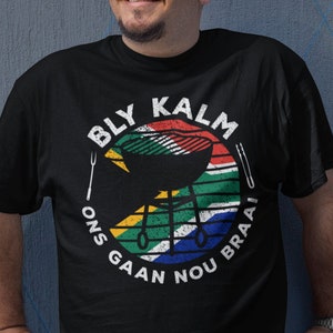 May include: A black t-shirt with a white circle graphic featuring a South African flag, a grill, and the text "Bly Kalm Ons Gaan Nou Braai".