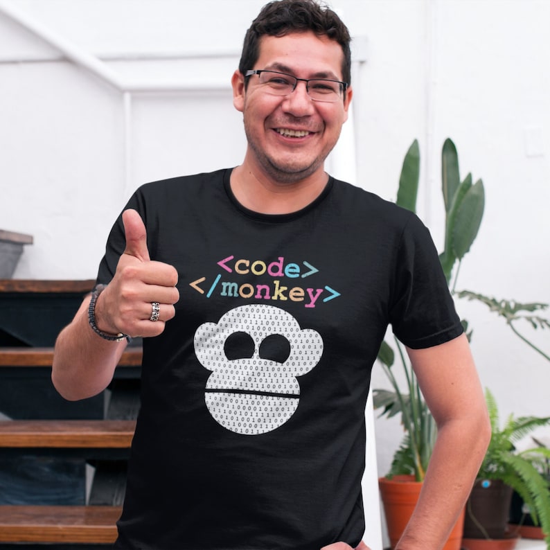 Może przedstawiać: Czarny t-shirt z białą małpią twarzą wykonaną z kodu binarnego. Tekst "<code>/monkey>" jest nadrukowany nad twarzą małpy.
